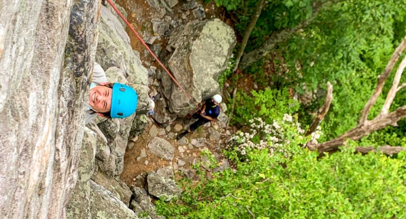 a girl who is rock climbing smiles up at the camera while a person belaying looks on from below
